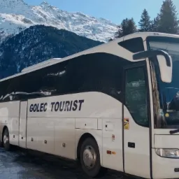 Autokar 'GOLEC TOURIST' na tle ośnieżonych gór i zabudowań. Biały pojazd z czarnymi akcentami, widoczny numer rejestracyjny. Zimowy krajobraz, słoneczny dzień.