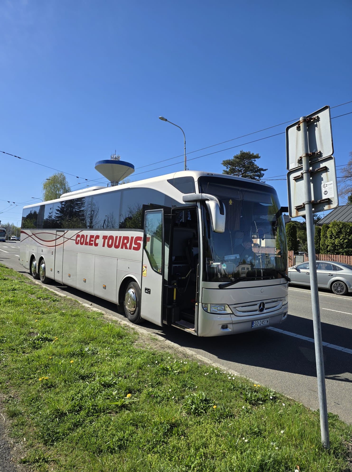 Nowoczesny, srebrny autokar turystyczny z otwartymi drzwiami na tle błękitnego nieba i zieleni. Widoczny znak firmowy 'GOLEC TOURIST'.