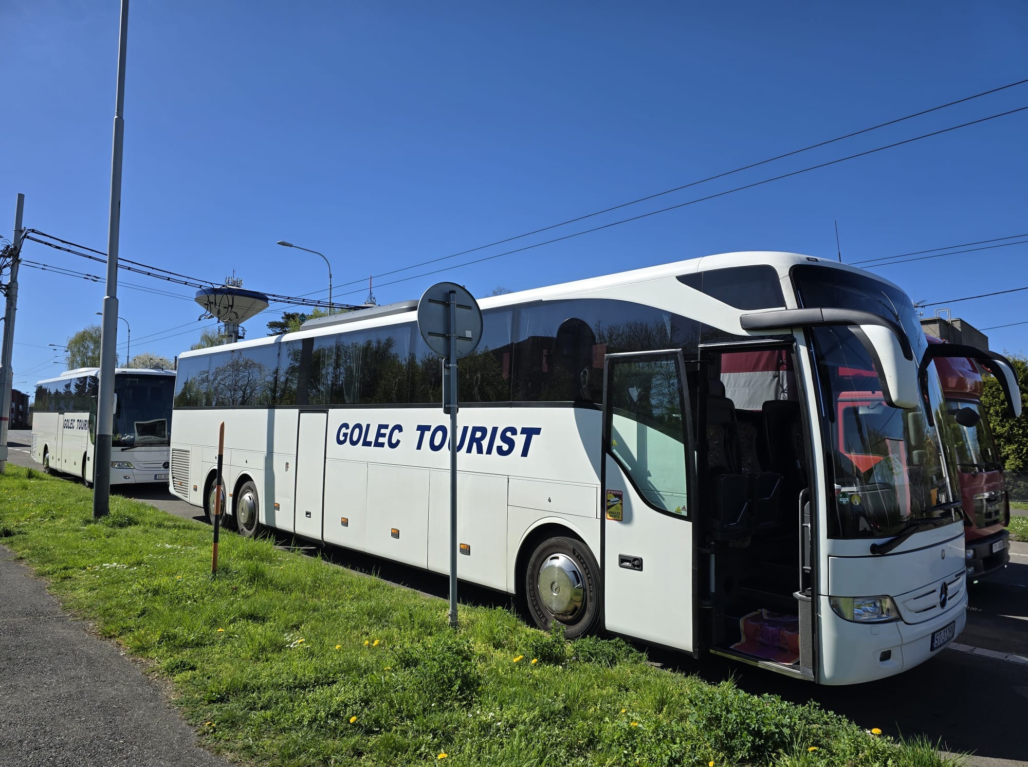 Biały autokar turystyczny z otwartymi drzwiami na tle zieleni i błękitnego nieba. Widoczny napis 'GOLEC TOURIST' i zaparkowane pojazdy obok.