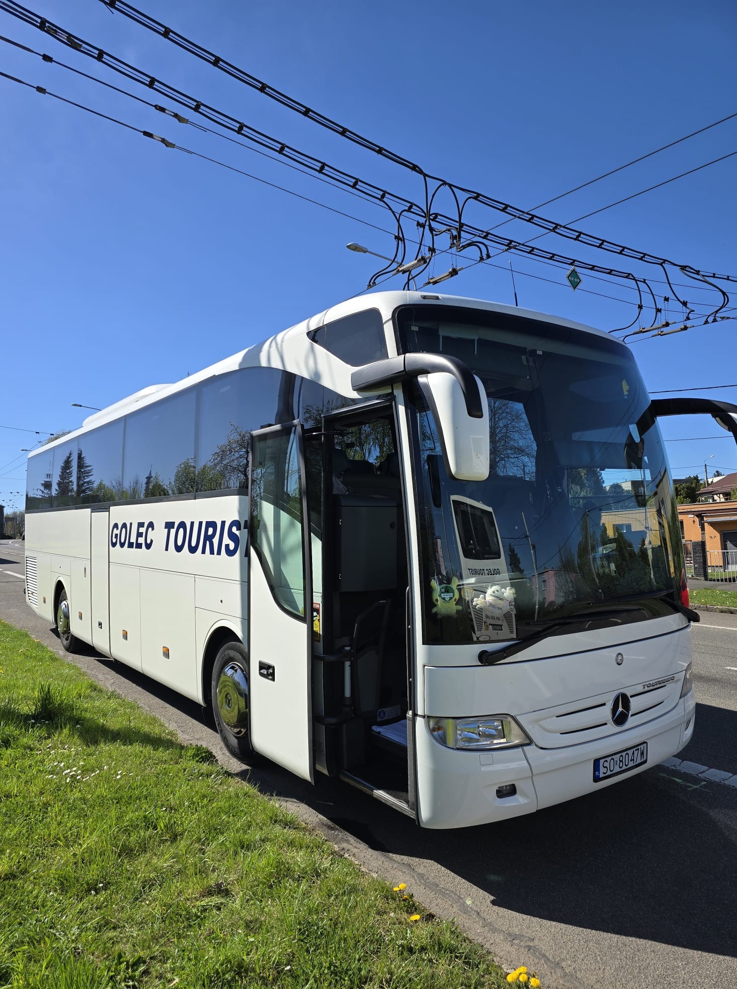 Biały autokar GOLEC TOURIST z otwartymi drzwiami stoi na poboczu drogi, widoczne linie trolejbusowe na tle błękitnego nieba. Tablica rejestracyjna SO 8047W.