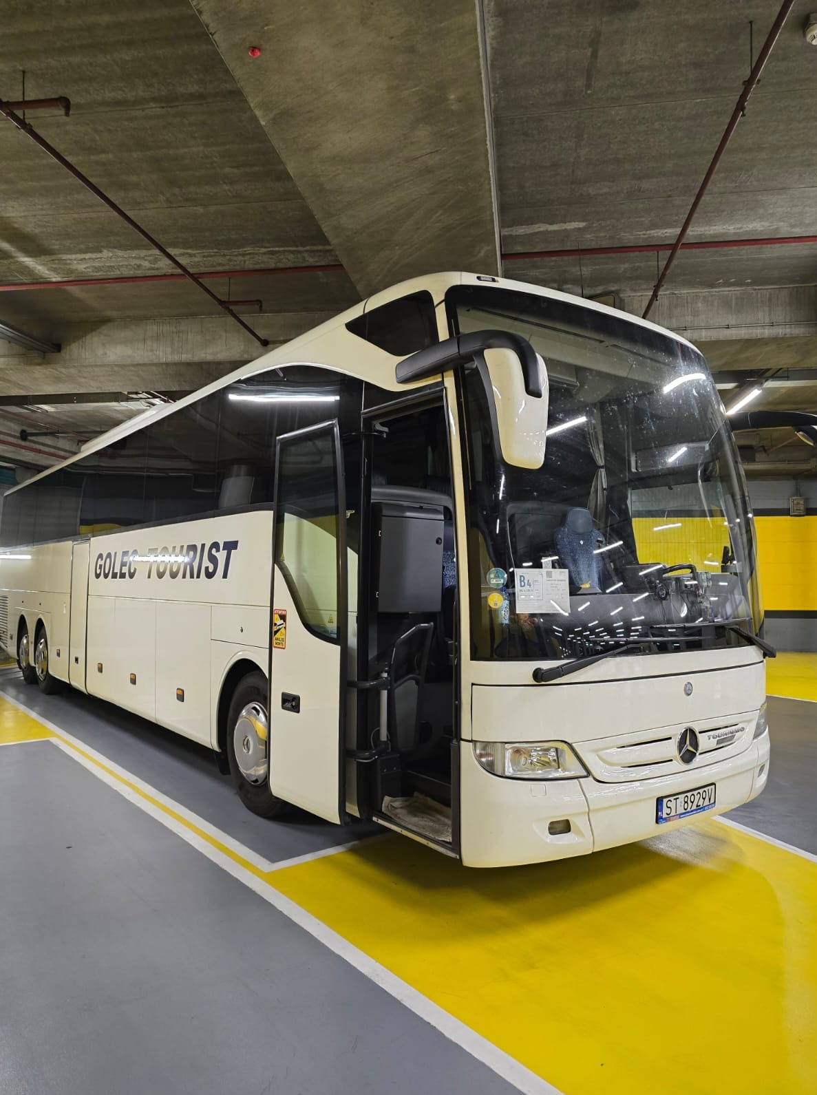 Autokar turystyczny marki Mercedes z otwartymi drzwiami, zaparkowany w garażu podziemnym z żółto-szarą nawierzchnią. Widoczny numer rejestracyjny.