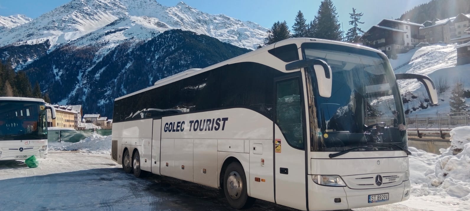 Autokar 'GOLEC TOURIST' na tle ośnieżonych gór i zabudowań. Biały pojazd z czarnymi akcentami, widoczny numer rejestracyjny. Zimowy krajobraz, słoneczny dzień.