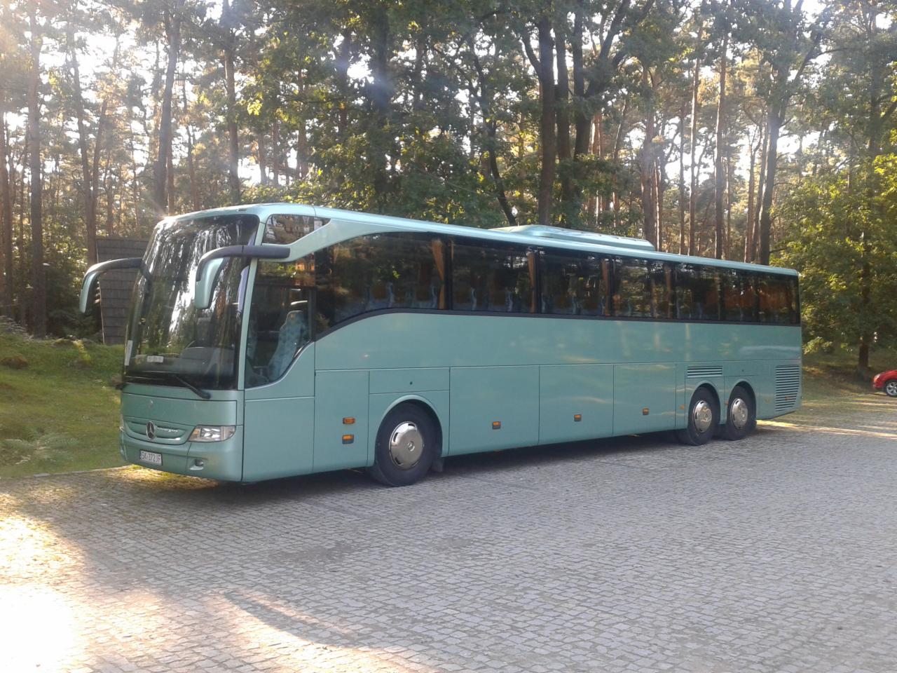 Turkusowy autokar Mercedes-Benz Tourismo zaparkowany na brukowanym placu, otoczony drzewami i wczesnym porannym słońcem.