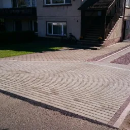 Ułożona kostka brukowa w odcieniach szarości i beżu tworząca podjazd i chodnik przed domem z dodatkowym pasem ozdobnego żwiru.