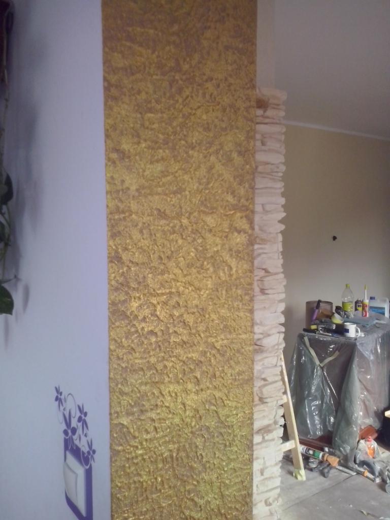 Złota ściana z fakturą, ozdobny tynk strukturalny na fragmencie ściany, obok imitacja kamienia. Widoczne narzędzia malarskie i zabezpieczenia.