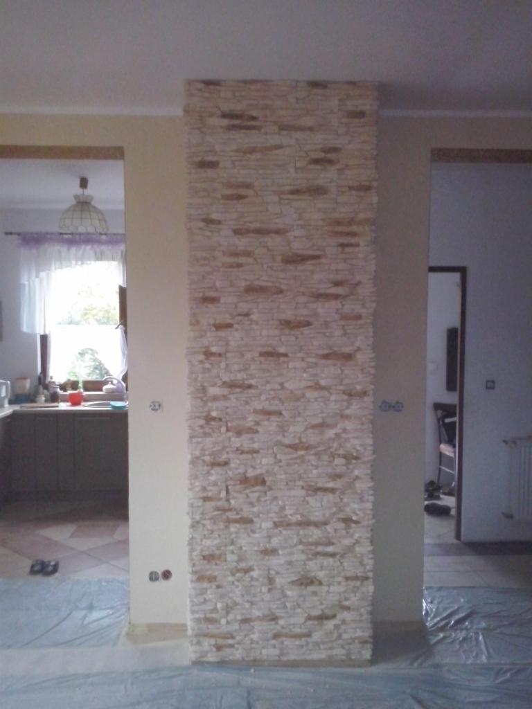 Dekoracyjny panel ścienny z jasnego kamienia na kolumnie, oddzielający dwa pomieszczenia. Podłoga zabezpieczona folią malarską. Widoczne gniazdka elektryczne.