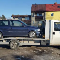 Niebieski Volkswagen Polo na platformie lawety, z budynkami w tle. Biała laweta z załadowanym autem gotowa do transportu na tle miejskiej zabudowy.