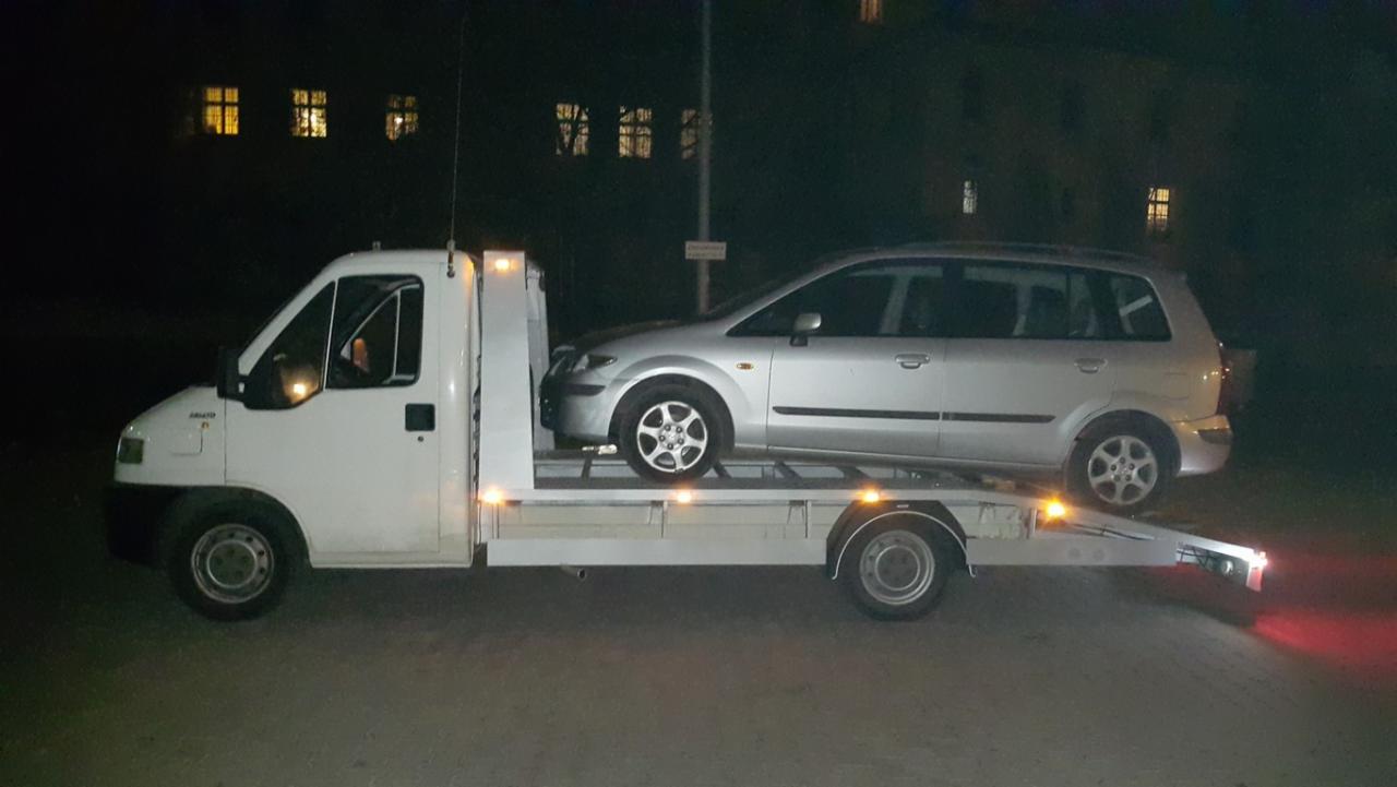 Biała laweta z srebrnym samochodem osobowym marki Mazda na platformie, nocą, oświetlona światłami pozycyjnymi, w tle budynek z oświetlonymi oknami.