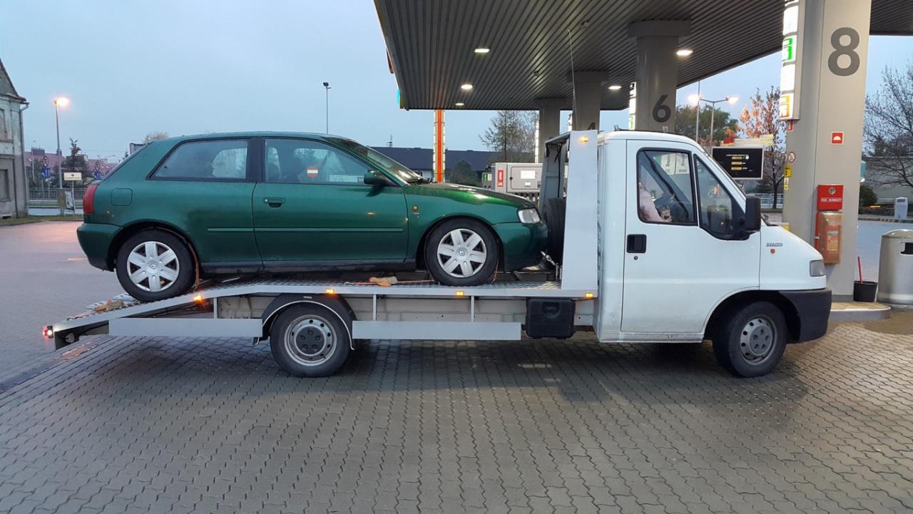 Zielony samochód osobowy na srebrnej lawecie przy stacji benzynowej w pochmurny dzień. Widoczny biały samochód lawety i dystrybutory paliwa.