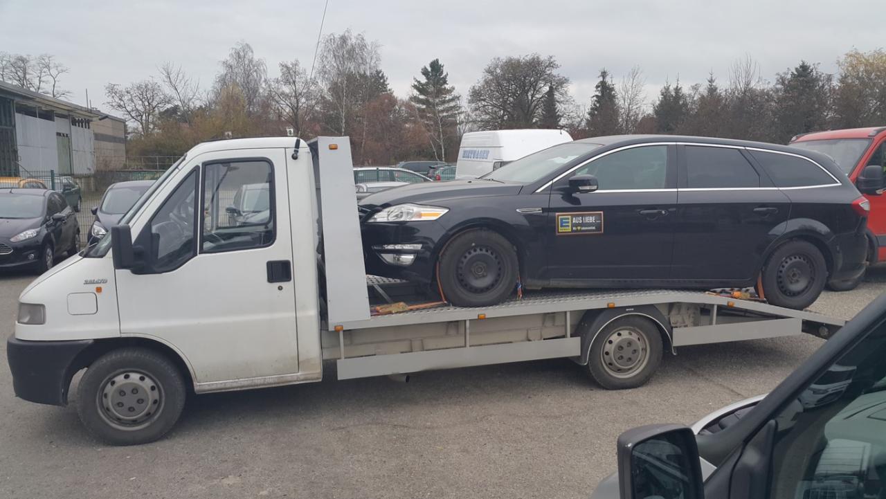 Czarny samochód osobowy na lawecie, zabezpieczony pasami transportowymi. Laweta w kolorze białym, widoczne budynki w tle i inne zaparkowane pojazdy.