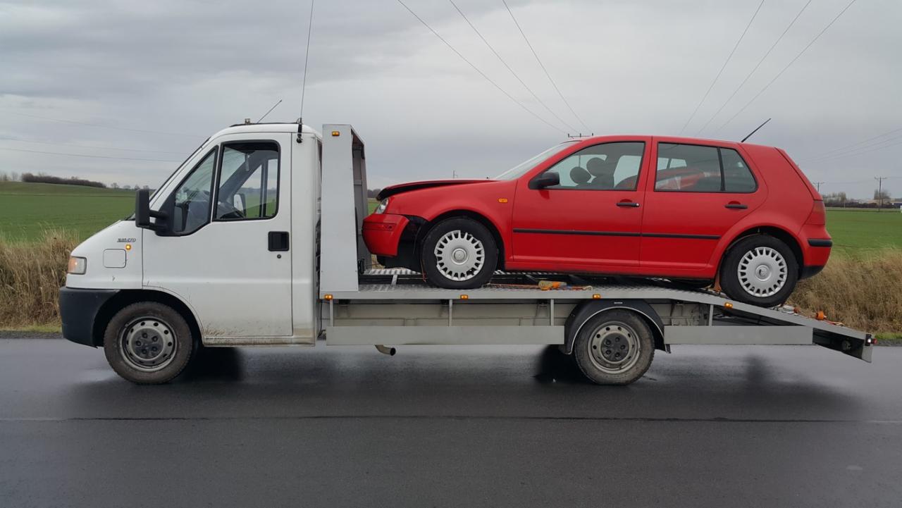 Biała laweta z uszkodzonym, czerwonym Volkswagenem Golfem na platformie, zaparkowana na mokrej drodze przy polu pod pochmurnym niebem.
