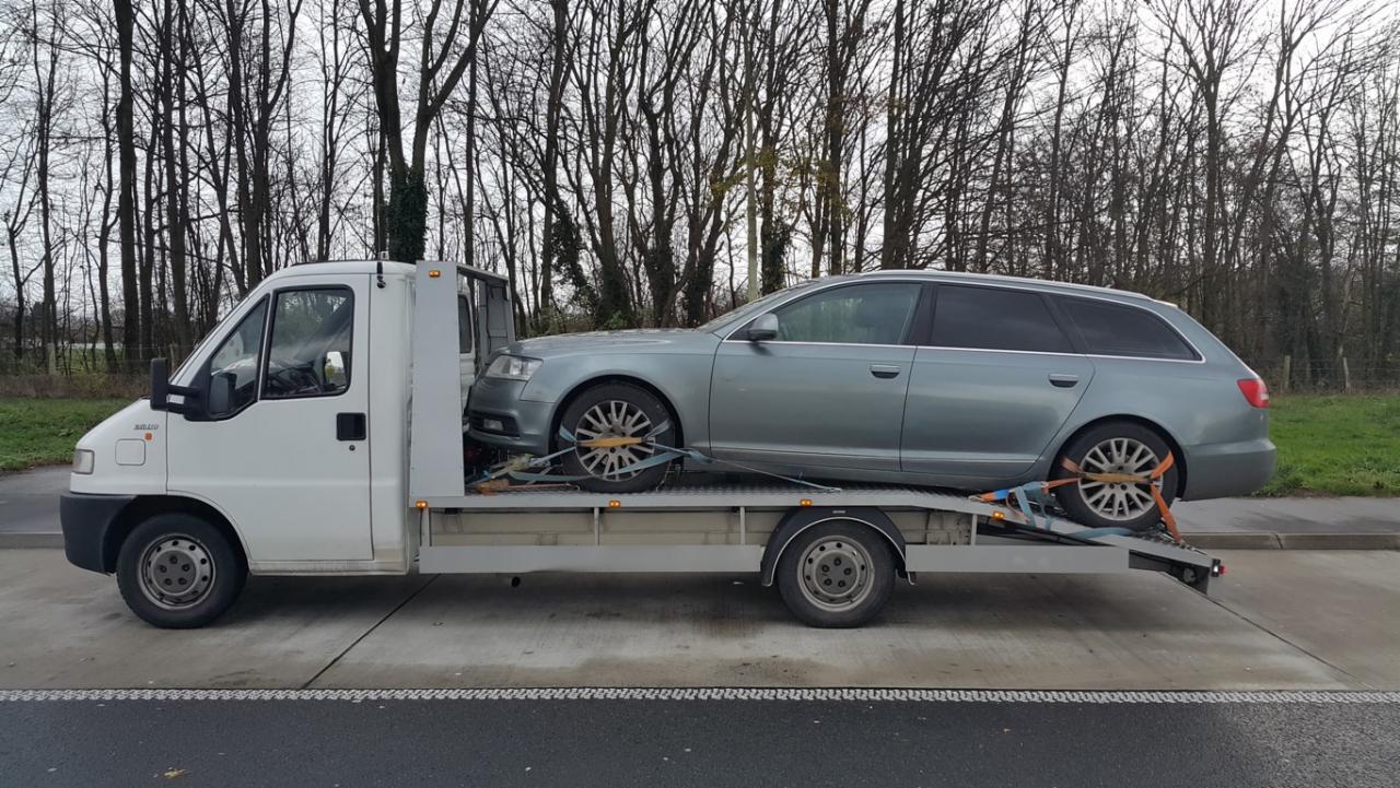 Laweta z szarym Audi kombi na platformie, zabezpieczonym pasami transportowymi. Biały samochód lawety na tle drzew bez liści i zieleni.