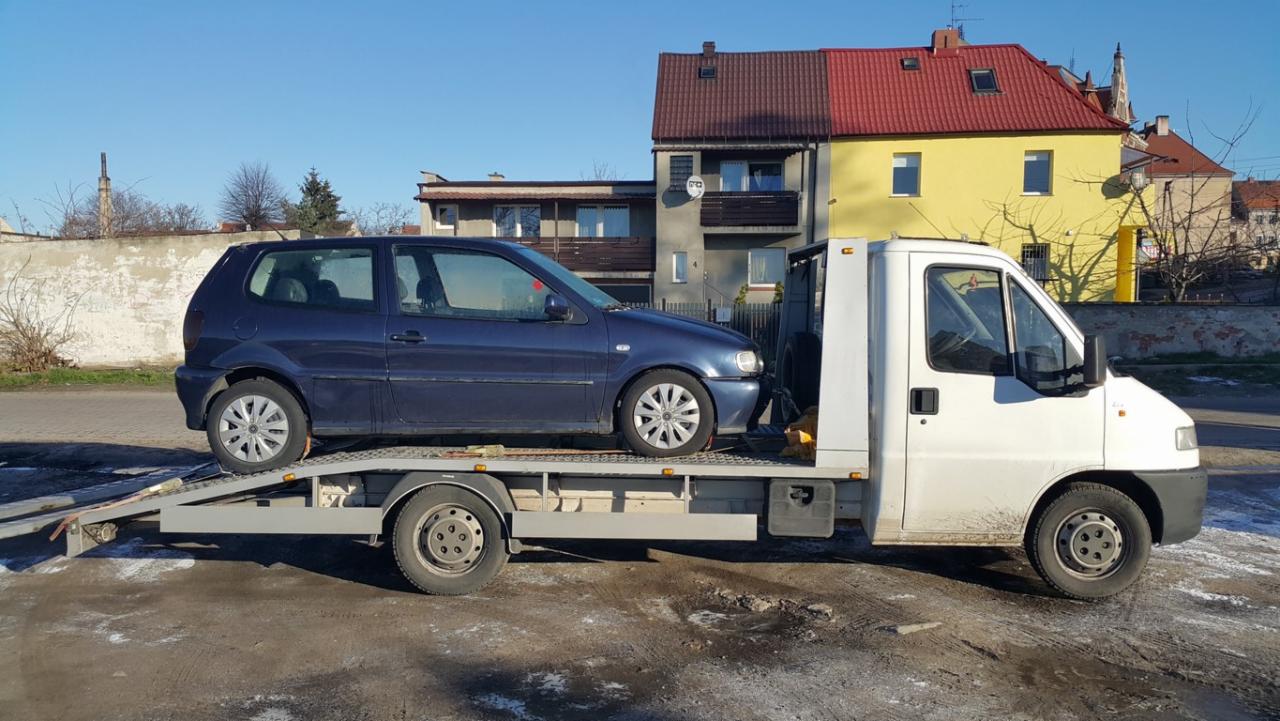 Niebieski Volkswagen Polo na platformie lawety, z budynkami w tle. Biała laweta z załadowanym autem gotowa do transportu na tle miejskiej zabudowy.