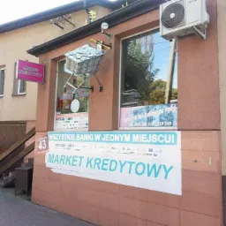 Fasada budynku z szyldem 'Market Kredytowy', w oknie wózek sklepowy wypełniony banknotami. Baner reklamowy z hasłem 'Wszystkie banki w jednym miejscu!'.