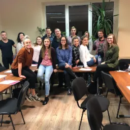 Grupa kilkunastu osób, siedzących i stojących za stołami w sali szkoleniowej, z butelkami wody i notatkami, w tle okno z rośliną doniczkową.