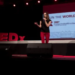 Prelegentka w okularach i czerwonych spodniach prezentuje na scenie TEDx Politechnika Wrocławska, mikrofon w dłoni, za nią ekran z napisem 'Run the World' i sylwetką budynku.