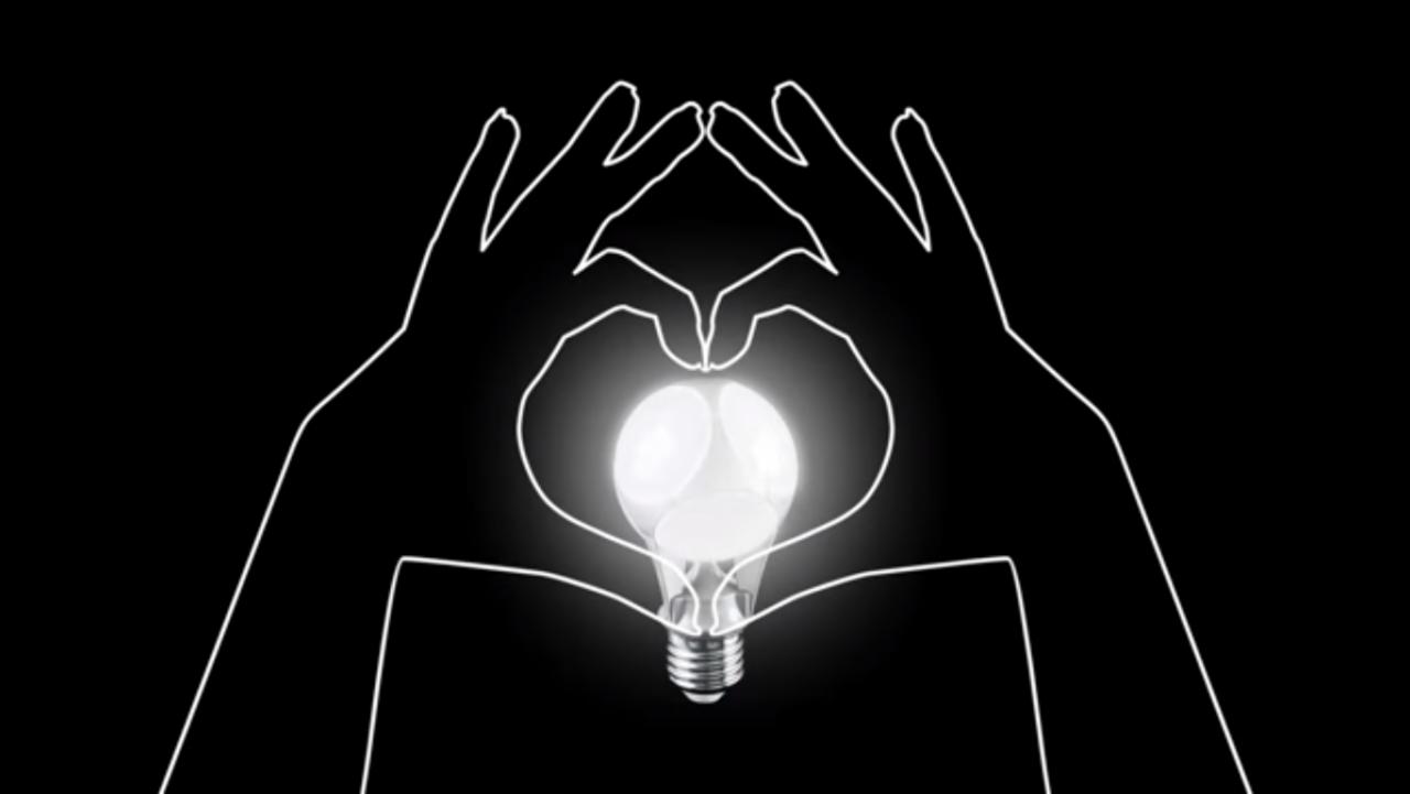 Stylizowana żarówka w dłoniach ułożonych w serce, na czarnym tle. Grafika w odcieniach bieli, symbolizująca pomysł lub kreatywność.