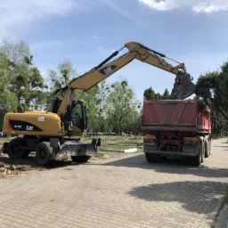 Żółta koparka CAT M316D ładuje ziemię do czerwonej ciężarówki na cmentarzu, w tle nagrobki i drzewa.