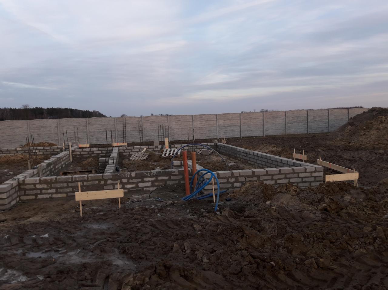 Wykop pod fundamenty domu murowanego z widocznymi zbrojeniami, częściowo wylane fundamenty z bloczków betonowych, rury instalacyjne i drewniane szalunki, w tle betonowe ogrodzenie panelowe.