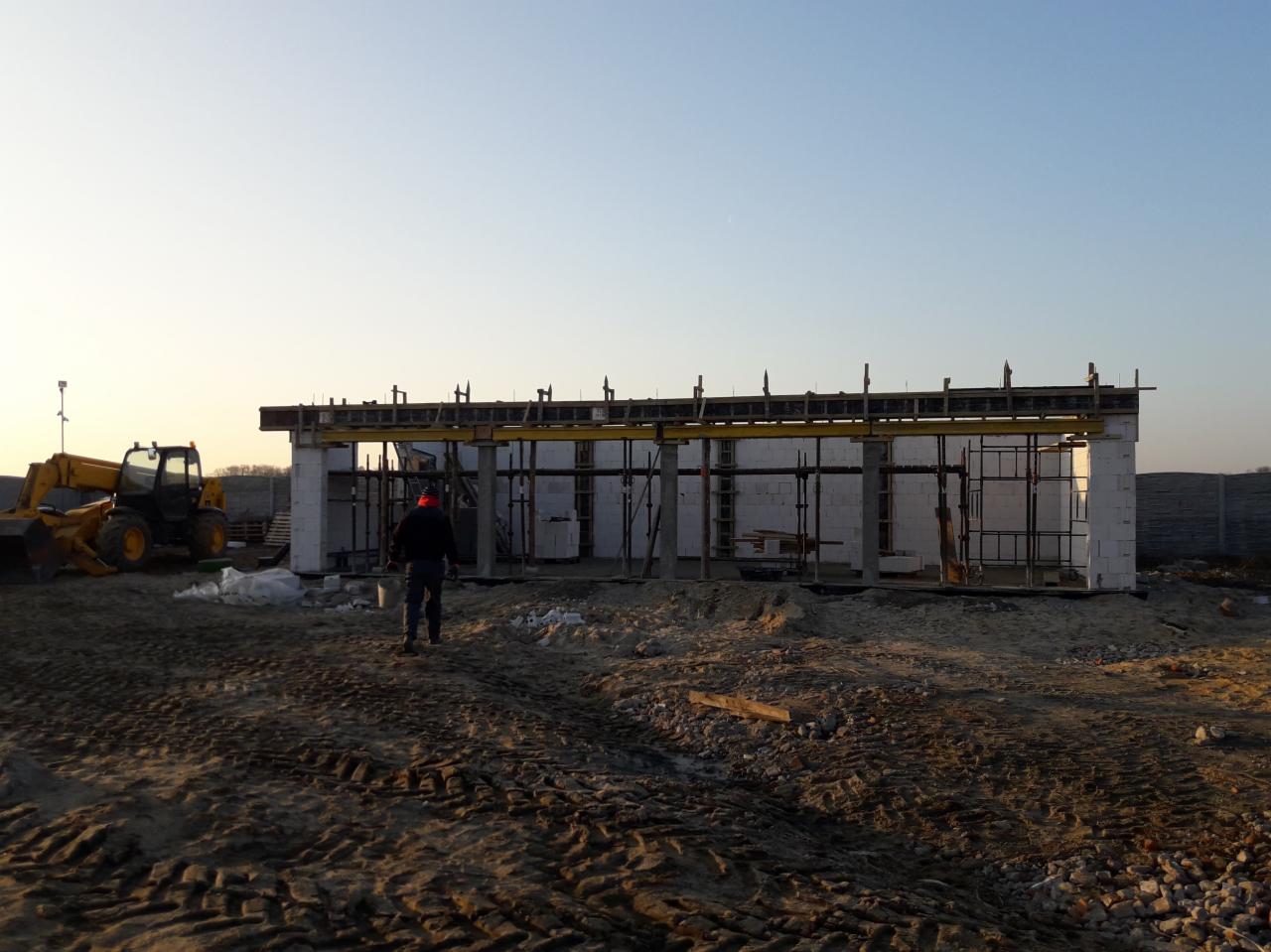 Budowa domu z betonu komórkowego w trakcie realizacji, widoczne szalunki i podpory, na pierwszym planie ślady opon koparki i pracownik w czerwonej czapce.