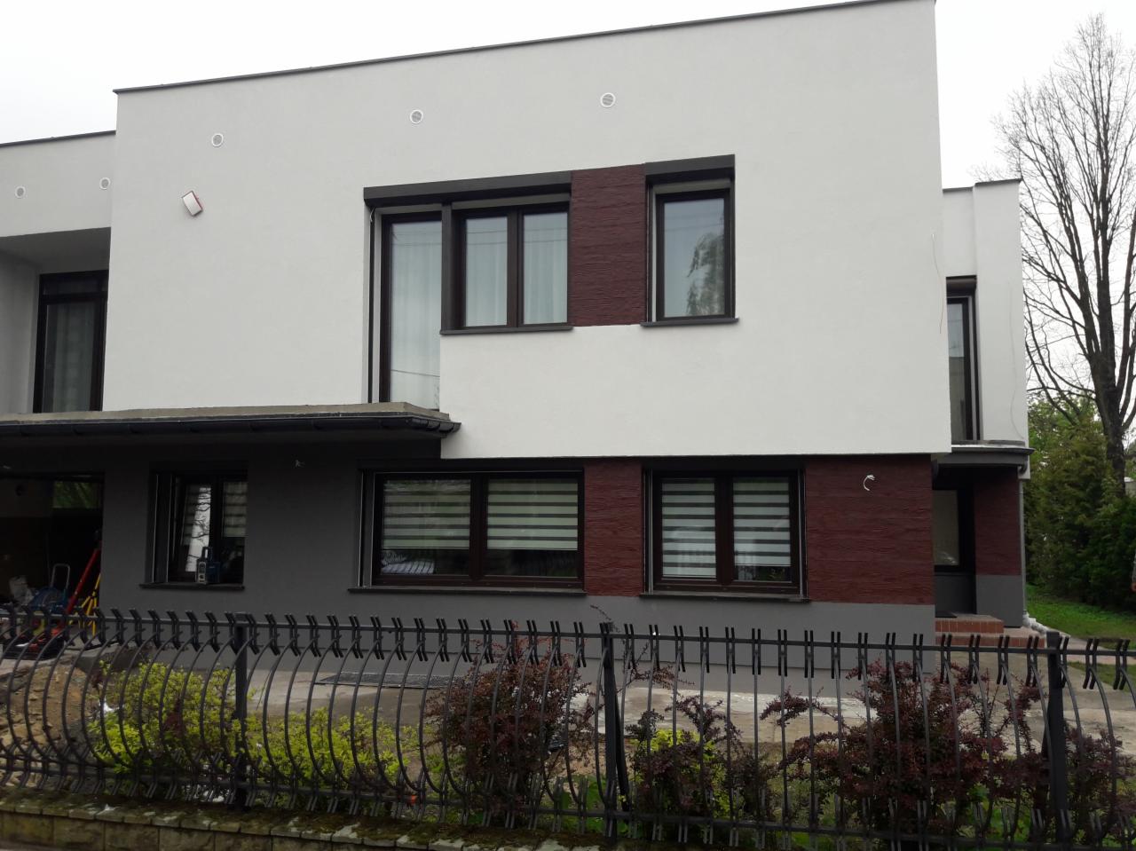 Dwukondygnacyjny dom z płaskim dachem, białą elewacją z brązowymi akcentami i czarną, metalową balustradą otaczającą posesję.