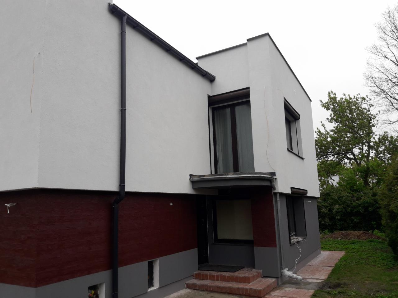 Dwukondygnacyjny dom z płaskim dachem, z pomalowaną na bordowo dolną częścią elewacji i białym tynkiem na górze, widoczne rynny i okna z ciemnymi ramami.