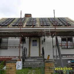 Dom jednorodzinny w trakcie wymiany pokrycia dachowego z użyciem rusztowania, widoczne ułożone dachówki karpiówki na łatach, komin z cegły i biała elewacja budynku.