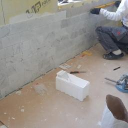 Bau - Układanie marmurowej mozaiki ściennej w trakcie prac wykończeniowych, widoczny glazurnik w stroju roboczym, narzędzia do cięcia płytek i pojemnik z zapasem mozaiki.