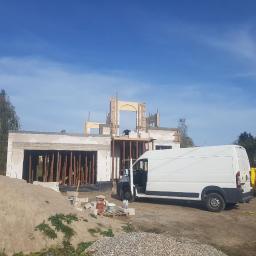 Bau - Budowa domu jednorodzinnego w stanie surowym otwartym, widoczna konstrukcja ścian z betonu komórkowego i drewniane szalunki otworów okiennych, biały samochód dostawczy na placu budowy oraz palety...