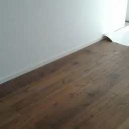 Ukończona podłoga z paneli imitujących ciemne drewno, jasne listwy przypodłogowe, biała ściana i fragment zabezpieczonej podłogi na dalszym planie.