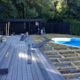 Budowa tarasu kompozytowego z widokiem na basen, widoczny szkielet konstrukcji i częściowo ułożone deski tarasowe w odcieniach szarości, w tle czarne ogrodzenie i budynek gospodarczy.