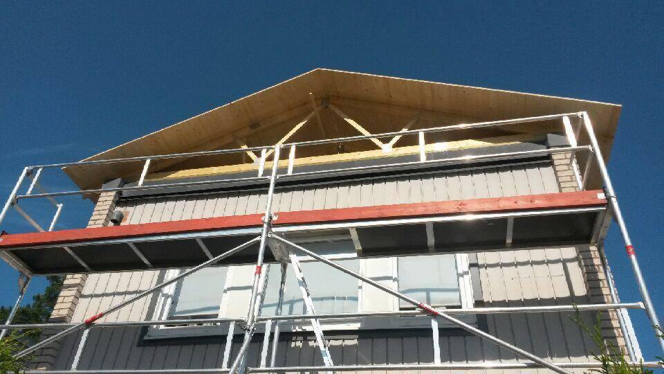 Dom w trakcie budowy z widocznym drewnianym szkieletem dachu i rusztowaniem przylegającym do ściany. Widoczne okna i fragment ściany z cegły.