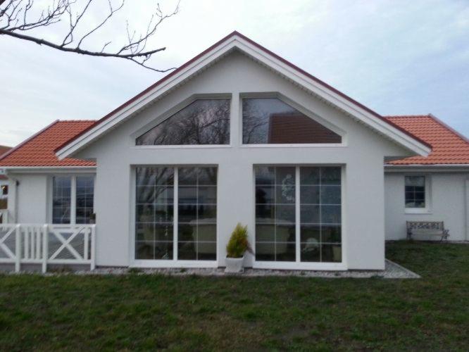 Biały dom z czerwonym dachem, dużymi oknami i trójkątnym oknem na poddaszu, otoczony trawnikiem i białym płotkiem.
