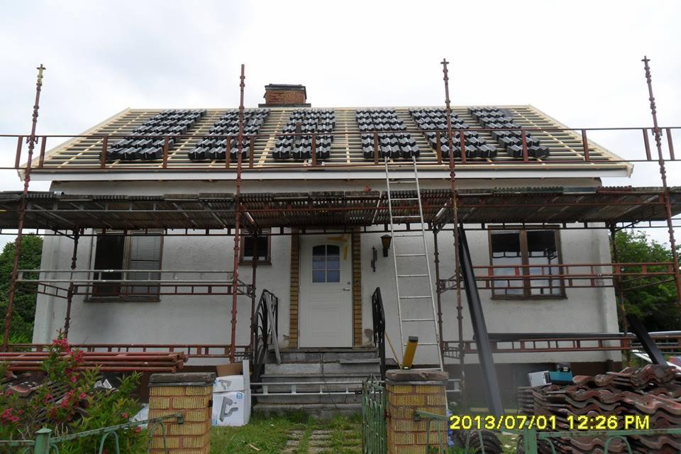 Dom jednorodzinny w trakcie wymiany pokrycia dachowego z użyciem rusztowania, widoczne ułożone dachówki karpiówki na łatach, komin z cegły i biała elewacja budynku.