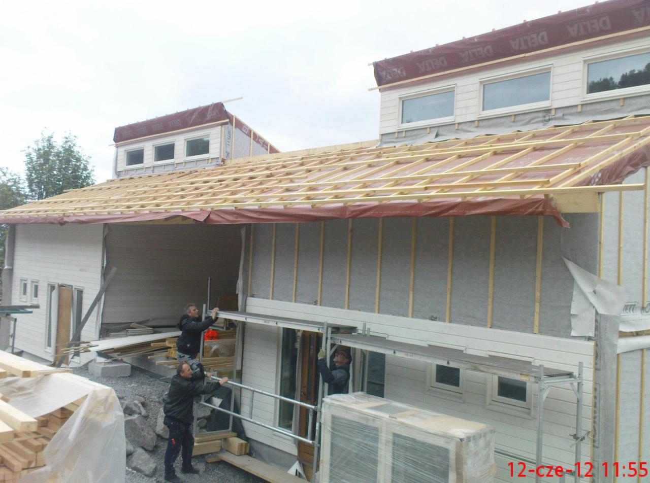 Dwóch pracowników montuje rusztowanie przy białym domu z drewnianą konstrukcją dachu w trakcie budowy, widoczne deski i materiały budowlane.