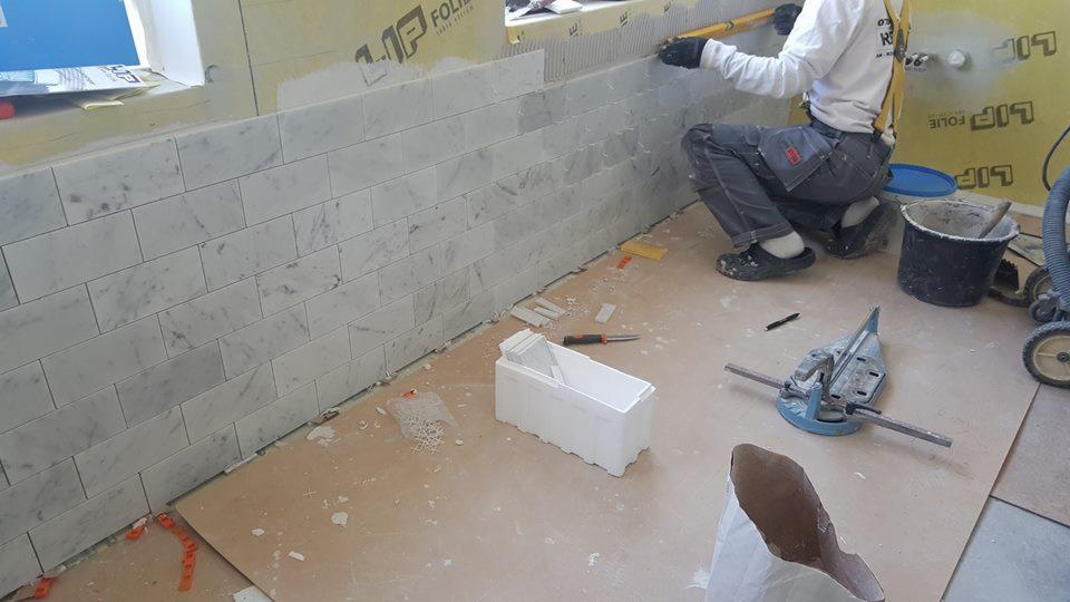 Układanie marmurowej mozaiki ściennej w trakcie prac wykończeniowych, widoczny glazurnik w stroju roboczym, narzędzia do cięcia płytek i pojemnik z zapasem mozaiki.