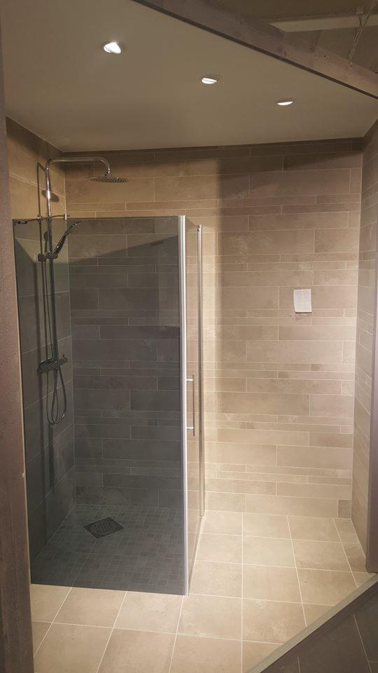 Nowoczesna kabina prysznicowa z deszczownicą i szklanymi drzwiami, wykończona płytkami w odcieniach beżu i szarości, z widocznym drewnianym elementem konstrukcyjnym pod sufitem.