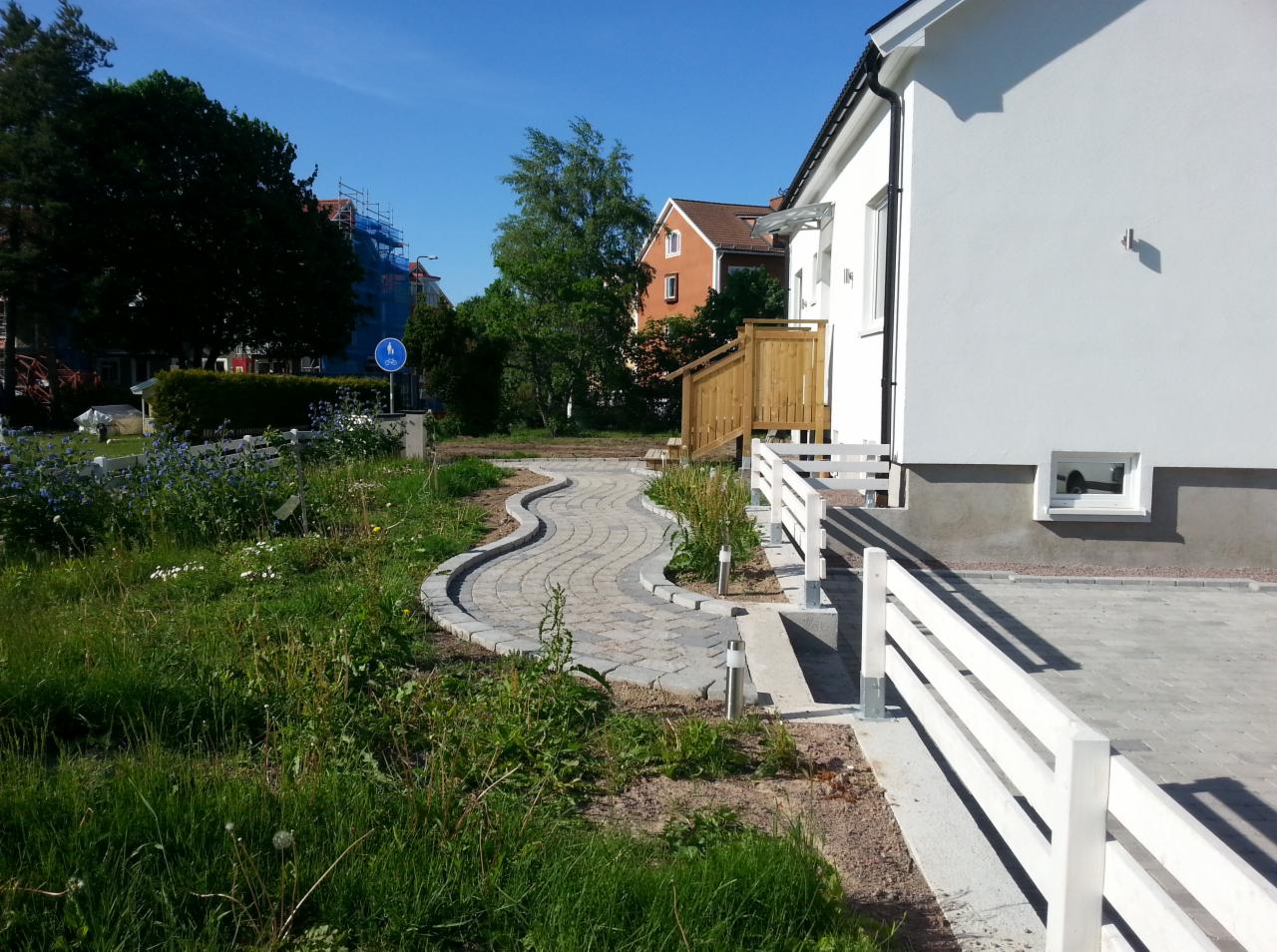 Ścieżka z kostki brukowej w kształcie litery S prowadząca do wejścia do białego domu z drewnianymi schodami i białą barierką, widoczny fragment posesji z zielenią i znak drogowy w tle.