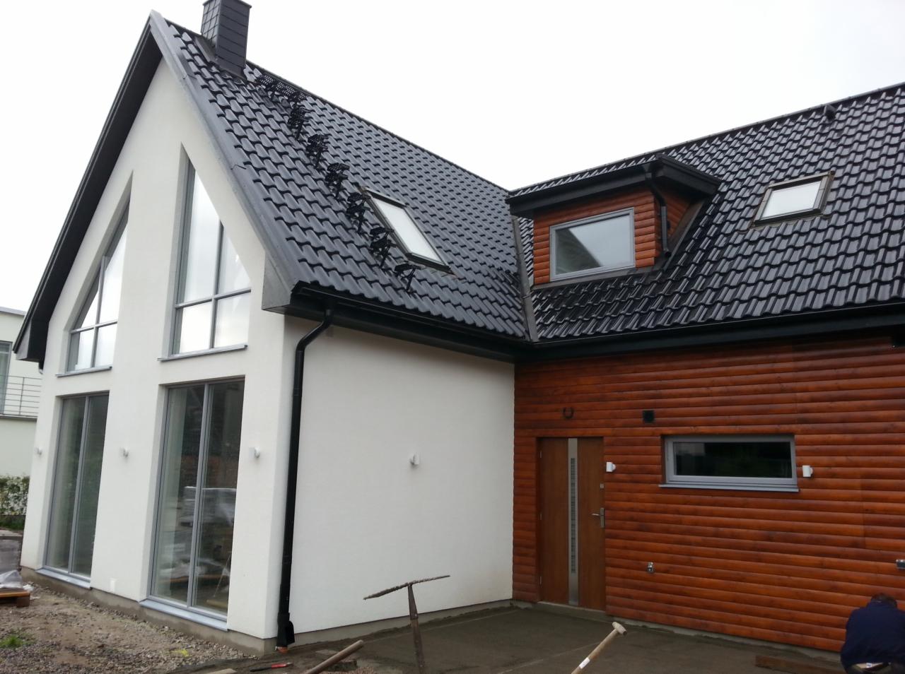 Nowoczesny dom z białym tynkiem i drewnianą elewacją, czarną dachówką i dużymi oknami, widoczne narzędzia budowlane sugerują prace wykończeniowe.