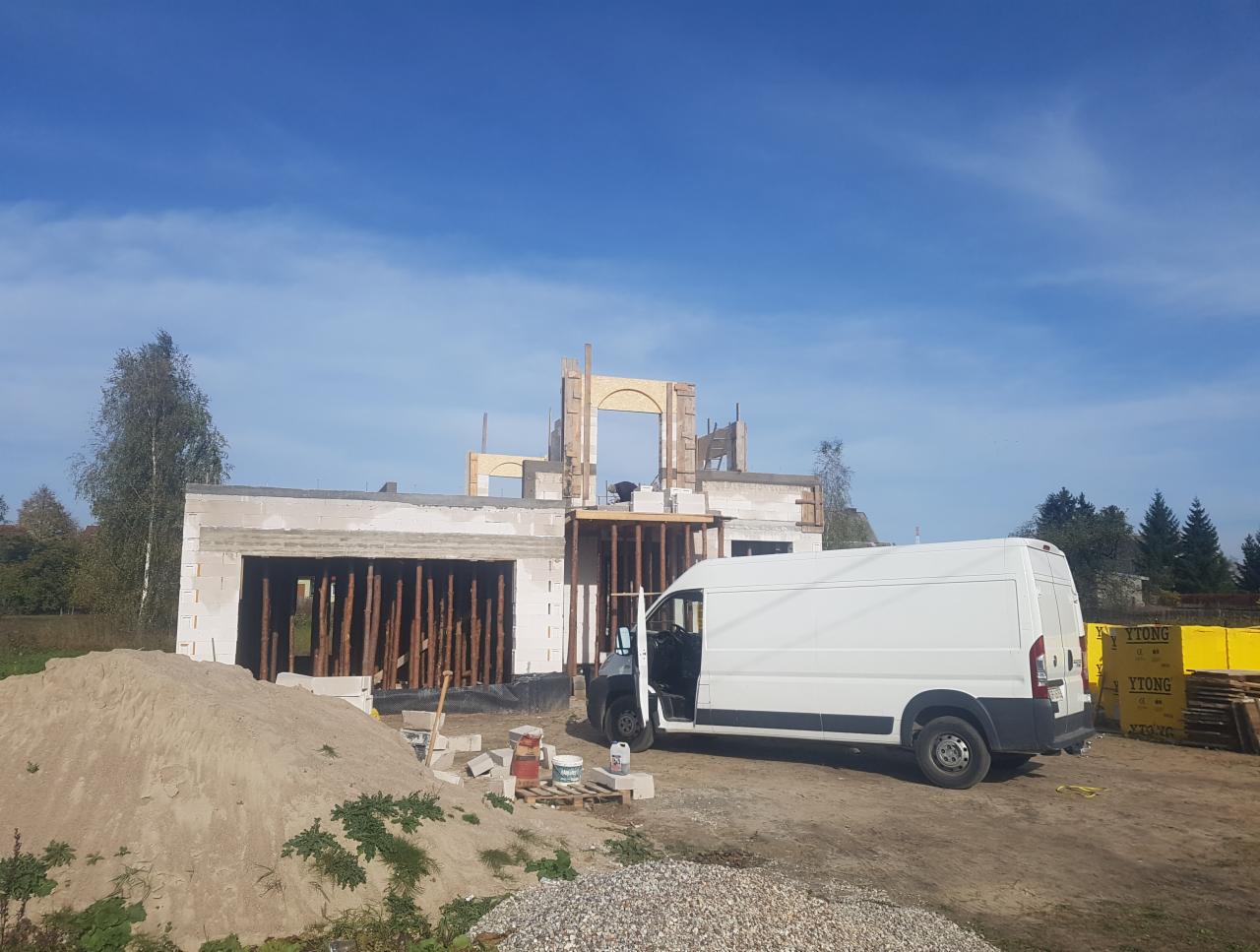 Budowa domu jednorodzinnego w stanie surowym otwartym, widoczna konstrukcja ścian z betonu komórkowego i drewniane szalunki otworów okiennych, biały samochód dostawczy na placu budowy oraz palety...