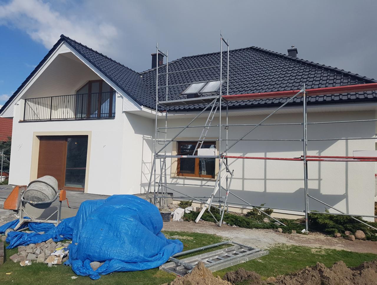 Biały dom jednorodzinny z ciemną dachówką, w trakcie prac wykończeniowych, z rusztowaniem przy elewacji i betoniarką oraz plandeką na trawniku.