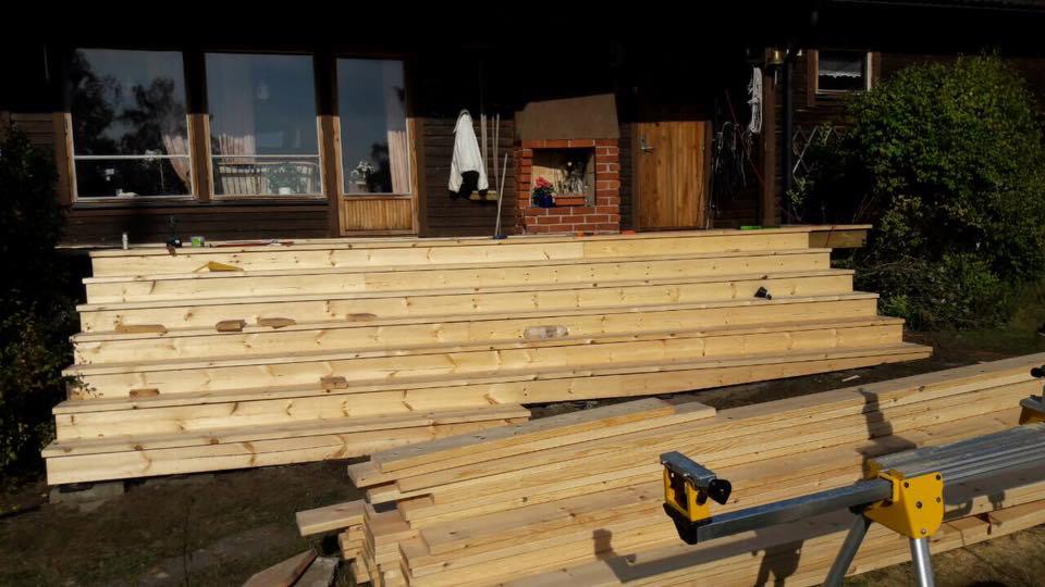 Konstrukcja drewnianych schodów wejściowych do domu z bali, widoczne deski i narzędzia, w tle okna i drzwi wejściowe.