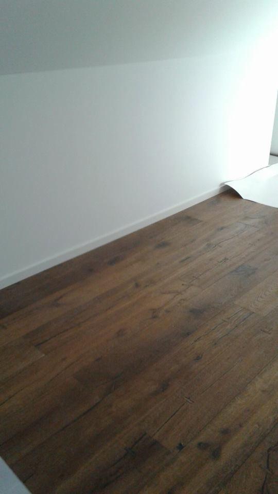 Ukończona podłoga z paneli imitujących ciemne drewno, jasne listwy przypodłogowe, biała ściana i fragment zabezpieczonej podłogi na dalszym planie.