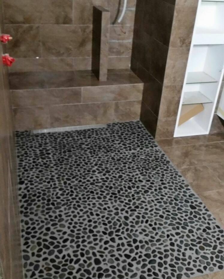 Prysznic z podłogą wyłożoną drobnymi, czarnymi kamieniami, brązowe płytki na ścianach i półka z białymi ramami.