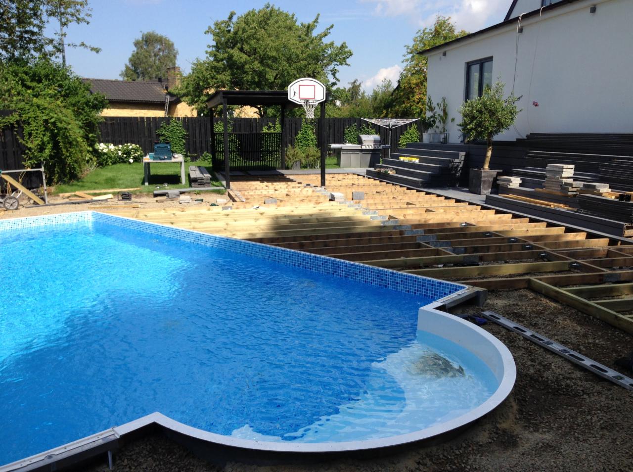 Budowa drewnianego tarasu wokół basenu w ogrodzie, widoczne elementy konstrukcyjne i deski tarasowe, w tle pergola, grill i kosz do koszykówki.