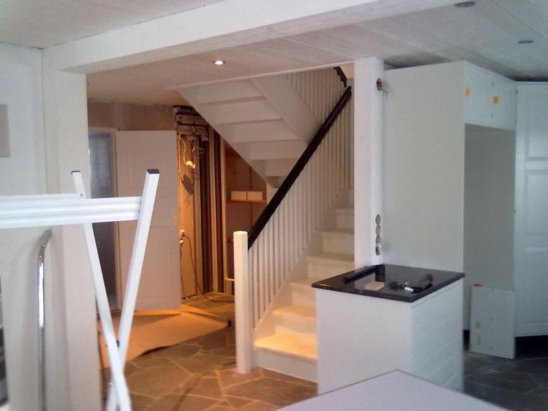 Wnętrze domu w trakcie remontu: widoczne schody z drewnianą poręczą, surowy stan ścian, fragment zabudowy kuchennej z czarnym blatem i podłoga wyłożona kamieniem. Część pomieszczenia jest w stanie...