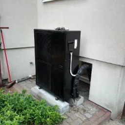 Czarna jednostka zewnętrzna urządzenia grzewczego na betonowym fundamencie przy ścianie budynku, widoczne rury w izolacji, para rękawic roboczych leży na górze obudowy.