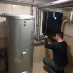 Instalator w trakcie podłączania czarnego kotła grzewczego Viessmann do srebrnego zasobnika ciepłej wody, widoczne rury z oznaczeniami przepływu.