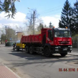 Czerwona ciężarówka Iveco z żółtym sprzętem i pracownikiem podczas naprawy drogi asfaltowej w słoneczny dzień. Widoczny znak stop i drzewa w tle.