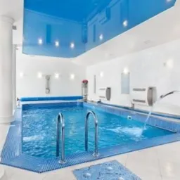 Błękitny basen wewnątrz budynku z kolumnami, wykończony mozaiką, z podświetlanym, błyszczącym sufitem napinanym i fontanną w tle.